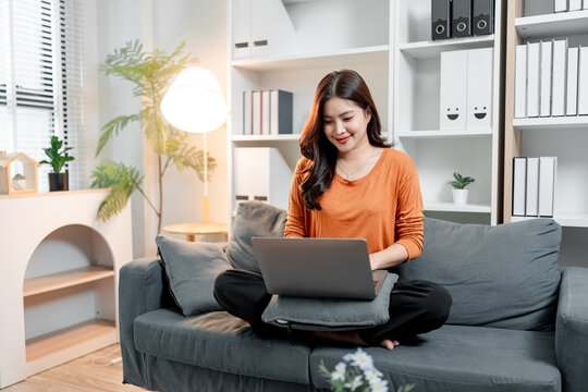 Young asian woman sitting comfortably on a gray sofa, working or studying on a laptop at home, embracing remote work and comfortable living concepts