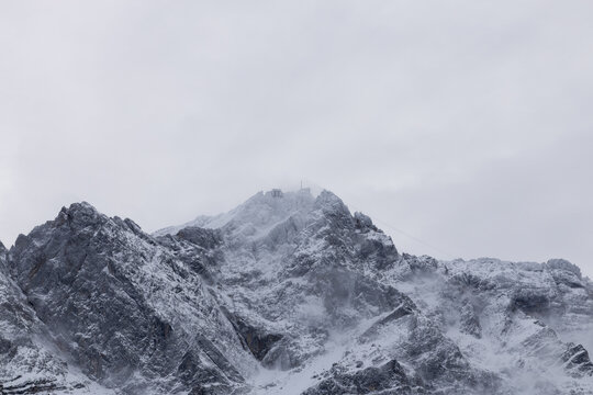 Majest&auml;tischer, schneebedeckter Berggipfel in den Alpen, umgeben von dichten Wolken und Nebelschwaden. Dramatische Winterlandschaft mit Felsformationen bei bew&ouml;lktem Himmel.