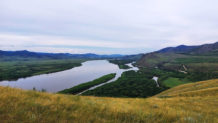 Fototapeta premium Winding Selenga River flowing through lush green valleys and forested islands in Buryatia, Siberia, with rolling hills under an overcast sky