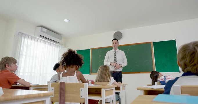 Male primary teacher leads math lesson from front of modern classroom while diverse elementary pupil group watches from desks, opening day routine supports attention, respect, academic readiness