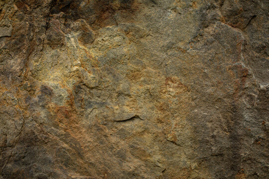 Raw, weathered rock face showing a rugged, uneven surface with natural cracks and chips. Rich organic color palette blending deep grey, rustic brown, ochre and golden hues.