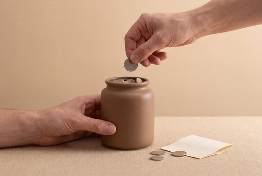 Hand dropping a coin into a ceramic donation jar beside loose coins and an envelope, charitable giving