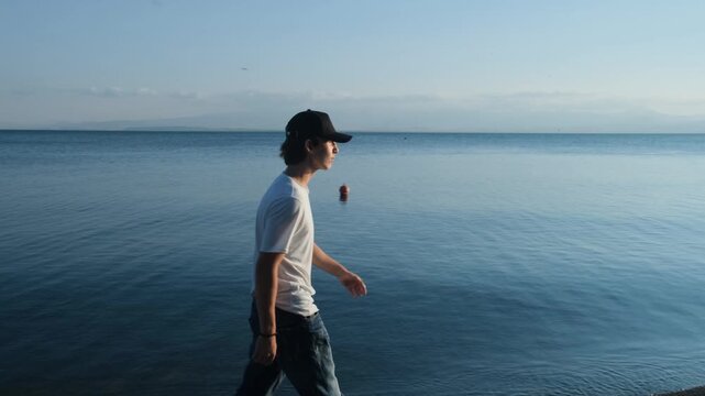Attractive young man in white T shirt, shorts and cap walks confidently along the shoreline at sunset. Model walk, beach sunset, self confidence and success mindset
