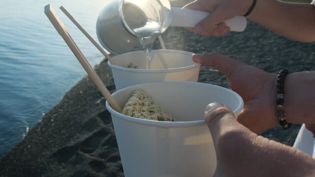 Pouring boiling water from a kettle into two portions of instant noodles on the seashore. Beach snack, hot food in paper plates, biodegradable disposable tableware and outdoor meal