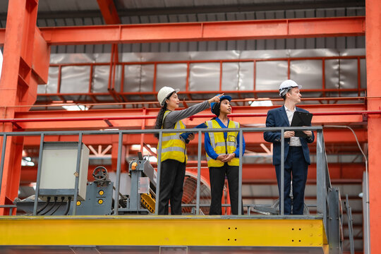 Female engineer points toward factory equipment while manager and coworker review production area, industrial inspection, manufacturing teamwork, operations management.