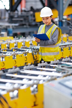 Female engineer records production information on clipboard beside industrial machinery, checking manufacturing equipment, quality control, factory inspection.