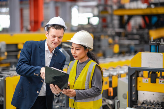 Factory manager explains production information to female engineer while reviewing clipboard beside industrial machinery, teamwork, inspection, quality control.