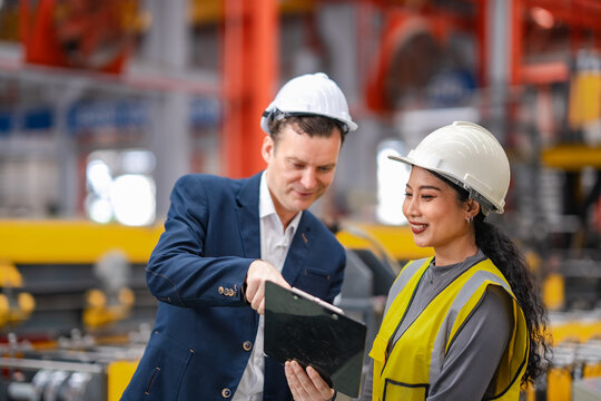 Factory manager and female engineer review clipboard together on production floor, discussing manufacturing process, machinery performance, quality control.