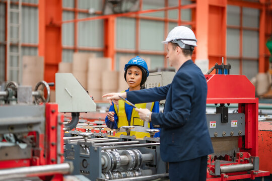 Factory manager and female engineer discuss production machinery on factory floor, reviewing industrial equipment, manufacturing process, teamwork, quality control.