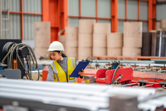 Female engineer checks production equipment while holding clipboard inside factory, monitoring industrial machinery, manufacturing operations, inspection, quality control.