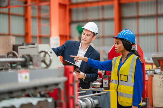 Female engineer points at industrial machinery while factory manager reviews equipment and clipboard, manufacturing inspection, production monitoring, teamwork.