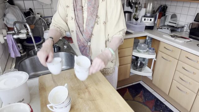 Woman begins sorting cups, plates and kitchen items from the sink onto a countertop before washing. Static Medium Shot.