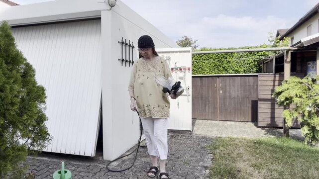 A woman removes dust from her clothing with compressed air after cleaning a vacuum cleaner container before setting the part down outdoors. Static Medium Shot.