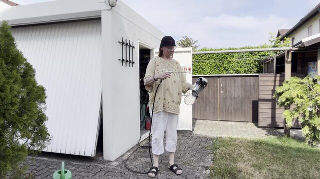 A woman uses compressed air to blow dust from a vacuum cleaner component outside an open garage in a sunny residential backyard. Static Medium Shot.