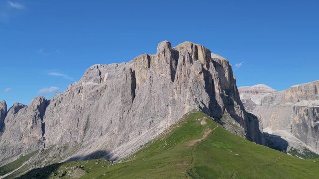 Aerial view of the rugged Sella Pass mountain range with green slopes and rocky peaks under a clear blue sky in Selva, Trentino-Alto AdigeS, Italy.