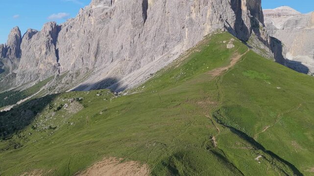 Aerial view of Sella Pass with green mountain slopes, rocky terrain, and winding hiking trails under bright daylight in Selva, Trentino-Alto Adige, Italy.