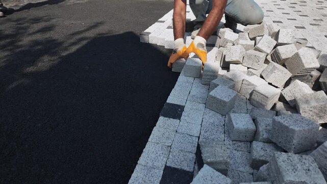 Wide angle view of worker hands installing interlocking concrete paving blocks on asphalt road during urban sidewalk construction and renovation project