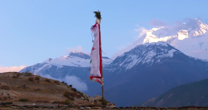 Annapurna Circuit, Manang Nepal, drone orbits around a vertical Buddhist prayer flag pole mounted on a stone base, revealing the snow-capped peak of Annapurna II in evening light.