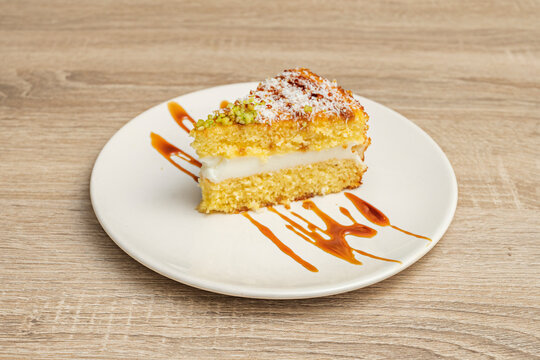 Layered sponge cake slice with cream filling, coconut, pistachio topping, and caramel drizzle on white plate