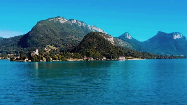 A breathtaking cinematic aerial reveal sweeping over majestic mountain ridges to expose the historic medieval Ch&acirc;teau de Duingt resting gracefully on a verdant peninsula jutting into Lake Annecy.