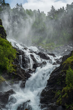 Vertical shot of white foaming water surging through dark rocks at L&aring;tefossen waterfall with dense water mist rising into forested mountain cliffs in Ullensvang, Vestland, Norway.
