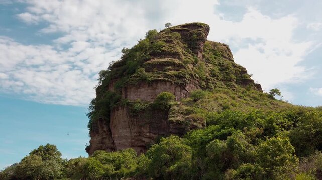 Scenic view of a massive layered rock formation covered with lush vegetation, rising above dense greenery beneath a blue sky with scattered clouds in Mexico
