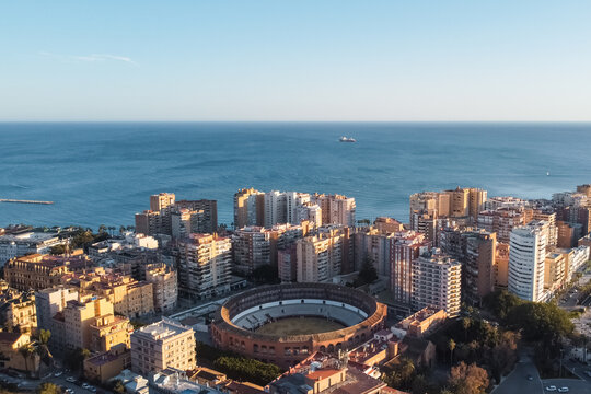 Aerial drone view of Malaga bullring beside Mediterranean coastline and urban skyline, Historic Plaza de Toros surrounded by Spanish city architecture and sea