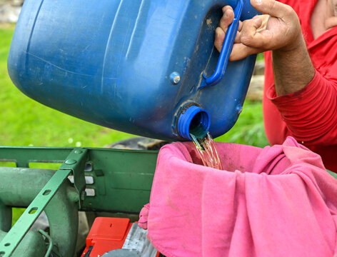 A person carefully pouring gasoline from a blue plastic fuel container through a cloth filter or strainer directly into a vehicle's fuel tank. A man pours fuel into a tractor.
