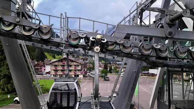Aerial view of a modern gondola cabin at the cable car station in the mountains with lush green trees and parking area in Alba, Trentino-Alto Adige, Italy.