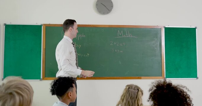 Male elementary teacher invites answer time near chalkboard as young girl walks forward and writes on board, supporting back to school participation, class discussion, and early math routine.