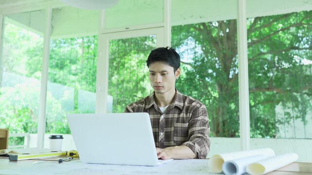 Architect working on building blueprints with a compass and laptop at a modern workspace, surrounded by rolled plans, measuring tools and color samples.