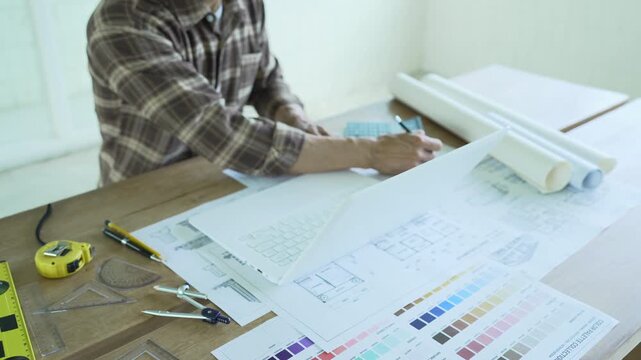 Architect drawing and reviewing detailed building blueprints on a desk with a laptop, rolled plans and color samples, representing architectural design and construction planning.