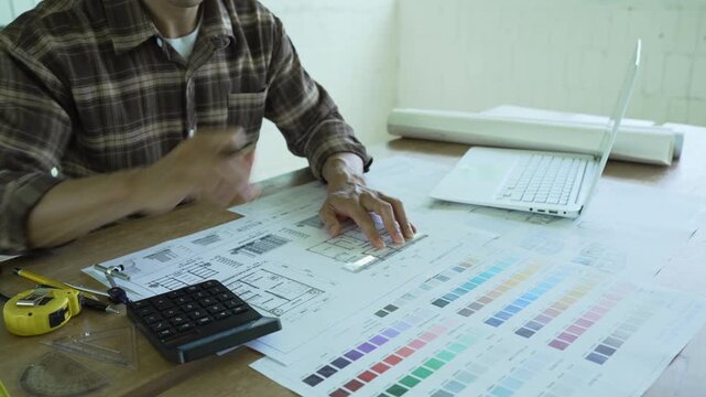 Architect drawing and reviewing detailed building blueprints on a desk with a laptop, rolled plans and color samples, representing architectural design and construction planning.