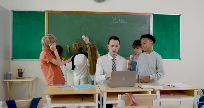 Male teacher works at laptop while diverse elementary pupils trade papers, talk, and give high five during group task, building classroom teamwork, friendship, motivation, and study routine.