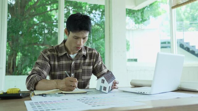 Architect working on building blueprints with a compass and laptop at a modern workspace, surrounded by rolled plans, measuring tools and color samples.