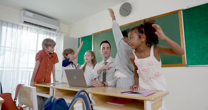 Wide classroom view follows young multicultural pupil group around male adult teacher as girl raises hand, joining question time, laptop activity, teamwork and back to school routine together