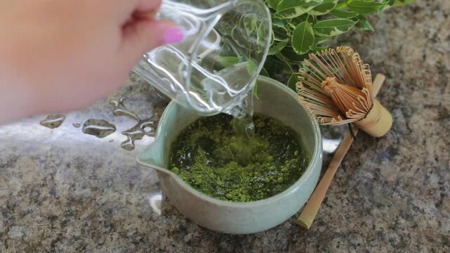 Pouring water into a small bowl of matcha powder, an essential step in preparing traditional matcha tea at home with a bamboo whisk nearby