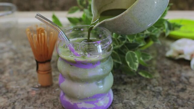 Pouring bright green matcha over purple ube milk in a glass, preparing a trendy homemade matcha ube latte at home