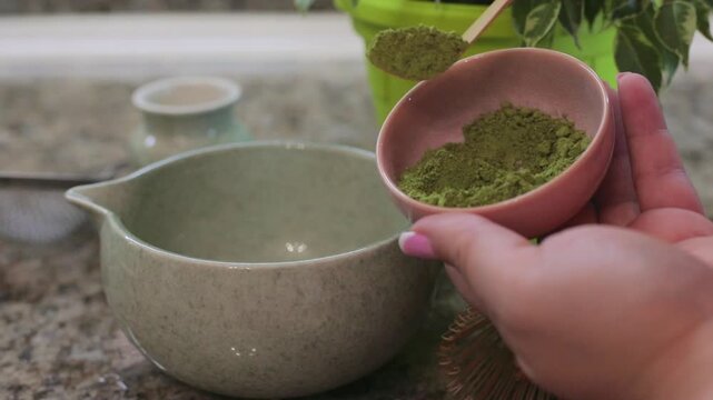 Scooping green matcha powder with a special matcha spoon into a ceramic bowl, preparing traditional matcha tea at home