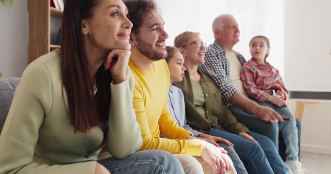 Family grandparents parents children watching tv smiling. Three generations relax on a sofa, sharing a cozy evening and following a show or live sports at home. Theme of happy family unity.