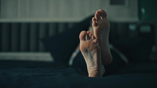 Low angle close up of female bare feet in sharp foreground with woman lying on bed and using smartphone in blurred bedroom background, cool moody light, relaxed home scene