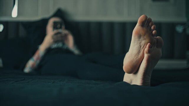 Low angle close up of female bare feet in sharp foreground with woman lying on bed and using smartphone in blurred bedroom background, cool moody light, relaxed home scene