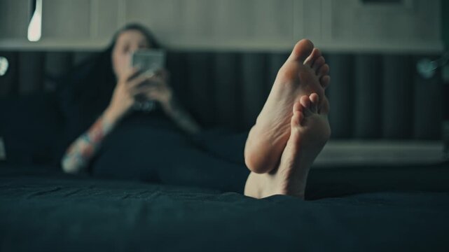Low angle close up of female bare feet in sharp foreground with woman lying on bed and using game console in blurred bedroom background, cool moody light, relaxed home scene
