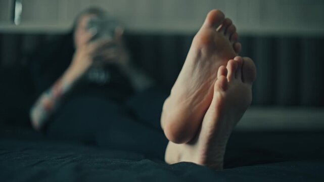 Low angle close up of female bare feet in sharp foreground with woman lying on bed and using game console in blurred bedroom background, cool moody light, relaxed home scene
