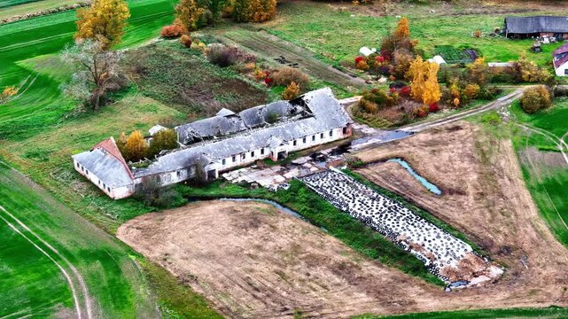 Aerial view of a dilapidated farm building with a damaged roof surrounded by agricultural fields and autumn trees in rural Latvia.