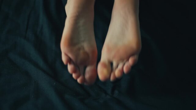 Close-up of female bare soles gently lifting and crossing on dark fabric with soft directional light revealing heel contours, toe pads, skin texture, and subtle repositioning