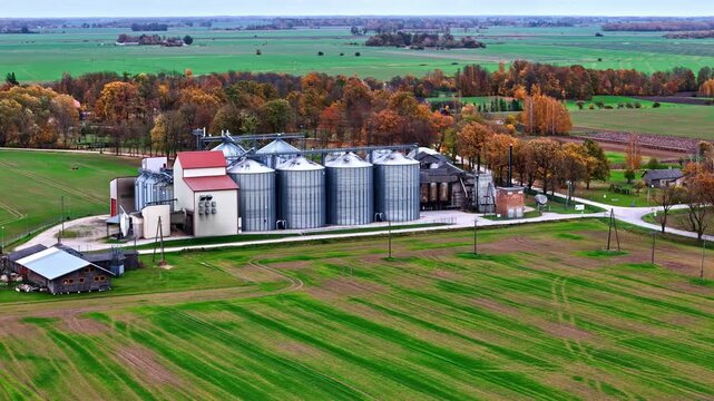 Aerial view of industrial grain silos surrounded by green fields and autumn trees in the countryside of Latvia.