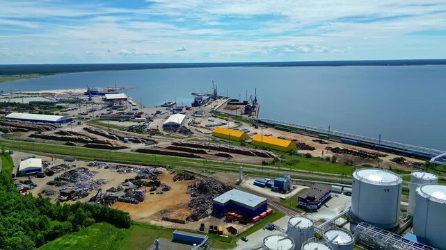 Aerial view of industrial port facility with storage tanks, timber piles, and cargo ships docked at the harbor in Latvia.