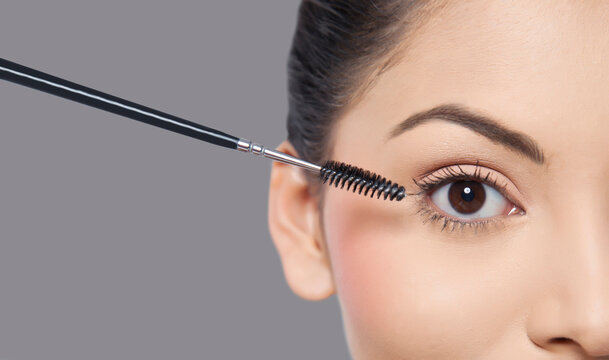 Young woman applying mascara over grey background 
