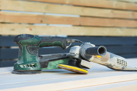 Electric orbital sander and angle grinder with sanding disc on wooden planks outdoors. Woodworking power tools, carpentry equipment or surface polishing concept for hardware store website banner.